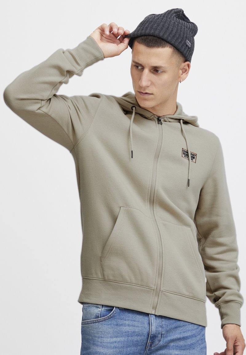 Sudadera con cierre en verde oliva claro, fabricada con un tejido suave. Cuenta con capucha, bolsillos frontales y un pequeño parche con logo en el pecho.
