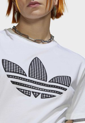Bílý bavlněný tričko s velkým černobílým kostkovaným logem Adidas trefoil, s černými vyšívanými konturami a horizontálními pruhy.