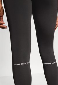 Leggings de sport noirs en tissu extensible. Présentent un texte blanc sur les jambes : "PROUVEZ-LES FAUX" et "NE JAMAIS ARRÊTER DE COMPÉTITIF".