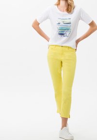 Camiseta blanca con un diseño texturizado en azul y verde en la parte frontal, combinada con jeans ajustados amarillos y zapatillas blancas.