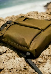 Olivgrüner, gepolsterter Rucksack aus strukturiertem Stoff, mit verstellbaren schwarzen Gurten und einem schlanken Design, der auf einer felsigen Oberfläche liegt.