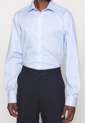 Homme portant une chemise habillée bleu clair à manches longues boutonnée, rentrée dans un pantalon bleu marine foncé, sur un fond uni.