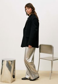 Veste noire oversize, pantalon large argenté brillant, et chaussures noires à motifs avec des cercles blancs. Reflets sur un tabouret métallique.