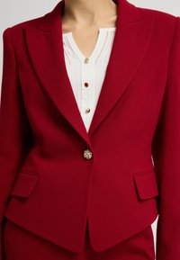 Červený saka na míru s výstřihem do V, strukturovanými rameny a jedním dekorativním knoflíkem. Spárováno s bílou plisovanou halenou, která má barevné knoflíky.