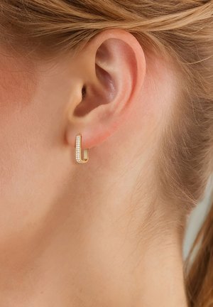 Gouden hoepeloorbel met een lineaire vorm, versierd met heldere crystals langs één kant. Draagt op een gepiercede oor.
