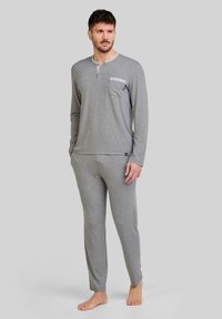 Hombre de pie descalzo, vistiendo una camiseta de manga larga color gris claro tipo henley con bolsillo en el pecho y pantalones a juego, contra un fondo liso.