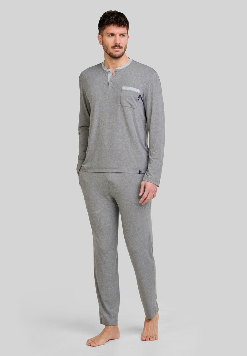 Hombre de pie descalzo, vistiendo una camiseta de manga larga color gris claro tipo henley con bolsillo en el pecho y pantalones a juego, contra un fondo liso.