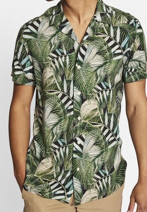 Homme portant une chemise boutonnée à manches courtes avec un imprimé de feuilles tropicales vertes et beiges, portée avec un pantalon marron clair.