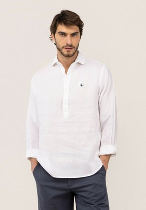 Hombre con cabello corto y castaño y barba, vistiendo una camisa blanca de manga larga con un pequeño logo y pantalones oscuros, manos en los bolsillos, fondo neutro.
