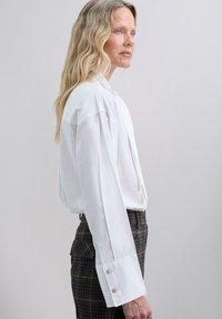 Blusa blanca de manga larga con un alto cuello, con un delantero plisado y puños oversized con botones decorativos. Combinada con pantalones de cuadro.