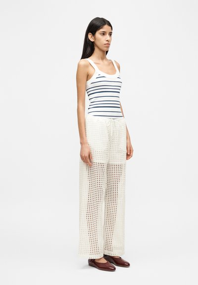 Ριγέ αμάνικο τοπ και ελαφρύ, διαφανές, με ανοιχτό ύφασμα παντελόνι σε εκρού χρώμα. Το μοντέλο φορά καφέ παπούτσια τύπου slip-on και στέκεται σε ουδέτερο φόντο.