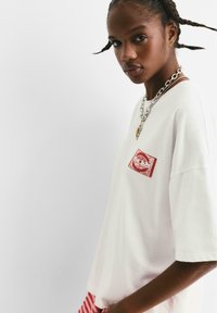 Ung kvinde med flettet hår iført en hvid oversized t-shirt med et rødt "Pacific" logo og lagdelte sølv kædeting.