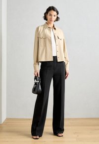 Beige zkrácená bunda s náprsními kapsami, černé široké kalhoty a černé sandály na pásky. Modelka drží malou černou kabelku.
