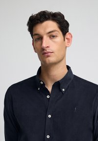 Camicia azzurra scuro con bottoni in tessuto morbido, caratterizzata da bottoni bianchi e un piccolo logo ricamato sul petto. Design con colletto button-down.