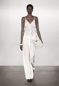 Witte, lichte gelaagde top met roesjes, gecombineerd met losse witte broeken. Accessoires omvatten een geweven tas en een chunky armband.