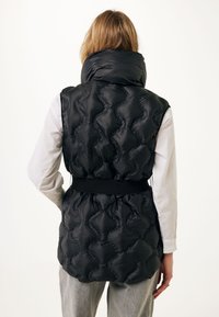 Černá péřová vesta s vysokým límcem, prošívaným vzorem, elastickým pasem a bočními kapsami, kombinovaná s bílou dlouhou rukávou košilí a šedými kalhotami.