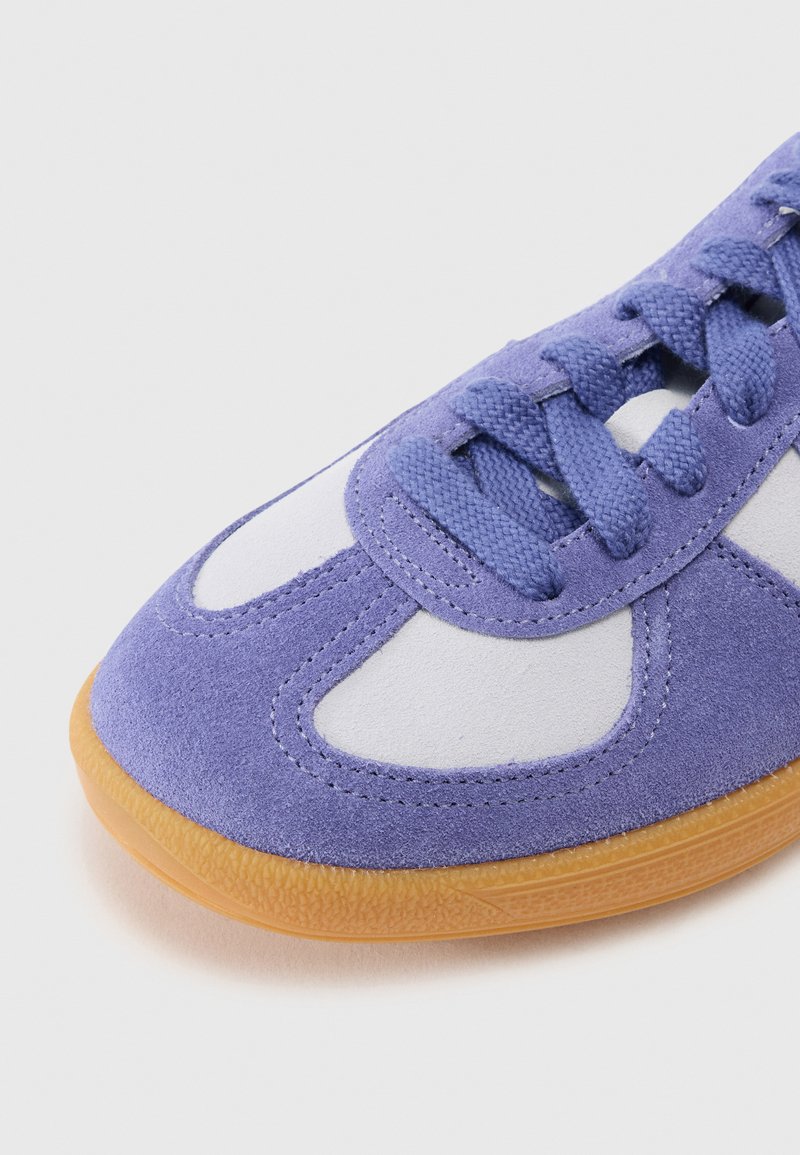 Sneaker viola in camoscio con dettagli bianchi, dotato di suola in gomma testurizzata, punta tonda e lacci in tessuto. Visione ravvicinata dei dettagli superiori.