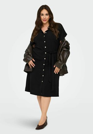 Žena s dugom tamnom kosom nosi crnu midi haljinu s gumbima i remenom, tamnu kožnu jaknu prebačenu preko ramena i tamne šiljaste cipele.