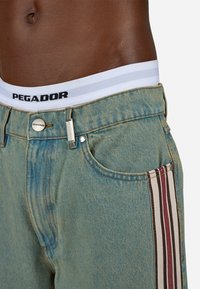 Svijetloplave traper šorts hlače s prugastim bočnim panelom u maroon i krem boji, s srebrnim gumbom i vidljivim logotipom brenda na pojasu.