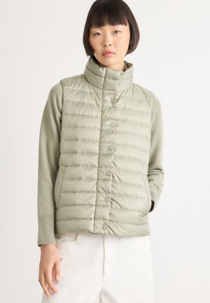 Femme jeune portant un gilet matelassé sans manches vert clair sur un pull à manches longues, se tenant avec une main dans la poche contre un fond blanc.