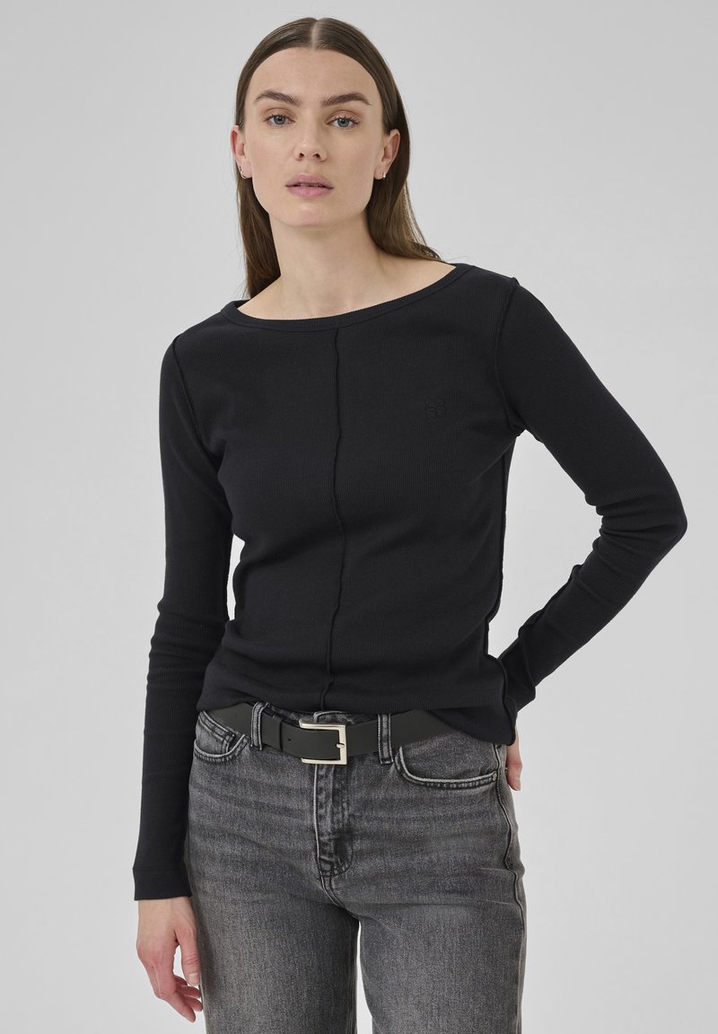 Woman wearing a black long-sleeve top and gray jeans with a black belt, standing against a plain light gray background.