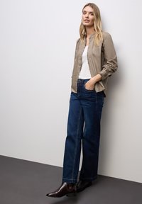 Camicia button-up marrone chiaro, canotta bianca, jeans a gamba larga blu scuro e stivaletti beige, esposti su uno sfondo neutro.