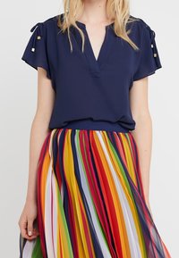 Femme portant un haut bleu marine à manches courtes avec des liens aux manches et une jupe plissée à rayures verticales aux couleurs de l'arc-en-ciel.