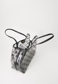 Przezroczysta torba typu tote w czarno-białe wzory, z czarnymi rączkami, charakteryzująca się miękkim, elastycznym designem i usztywnionym górnym otwarciem.