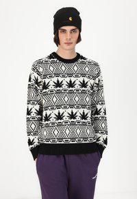 Black and white knitted sweater with a leaf pattern, round neck, and ribbed cuffs. Model wears a black beanie with a small logo and purple pants.