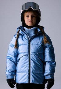 Mlada osoba s pletenom kosom nosi svijetloplavu jaknu s punjenjem, ski rukavice i kacigu s naočalama na jednostavnoj pozadini.