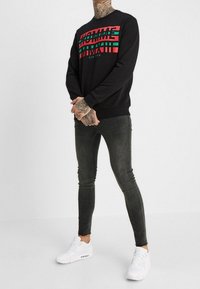 Sudadera negra con texto en negrita de rayas rojas y verdes, combinada con jeans oscuros ajustados y zapatillas blancas. Los detalles incluyen tatuajes en los brazos.