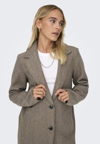 Manteau en mélange de laine marron avec un col en pointe, des boutons à l'avant et des poches latérales. Tissu légèrement texturé avec une coupe ajustée.