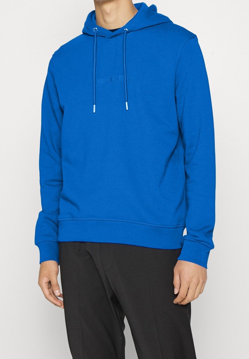 Uomo vestito con una felpa con cappuccio di un blu brillante e cordini, e pantaloni neri, in piedi contro uno sfondo semplice chiaro.