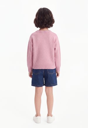 Enfant aux cheveux courts et bouclés portant un pull rose, un short en jean bleu et des baskets blanches, debout de dos sur un fond blanc.