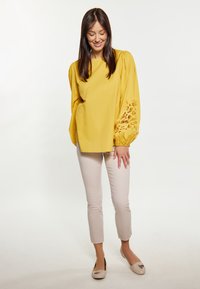 Blusa gialla con maniche a sbuffo e dettagli in pizzo ricamati, abbinata a pantaloni aderenti beige chiaro e scarpe flat beige.