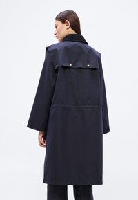 Μπουφάν τύπου τραντς σε ναυτικό μπλε χρώμα, ελαφρύ, με ψηλό γιακά, διακοσμητικά κουμπιά με κουμπώματα και χαλαρή εφαρμογή, που διαθέτει λεπτομέρεια με φάκελο στο πίσω μέρος.