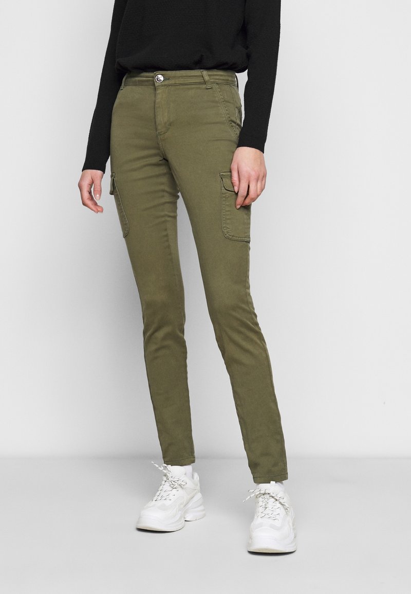 Femme portant un pantalon cargo slim olive avec poches latérales, un haut noir à manches longues et des baskets blanches, debout devant un fond uni.