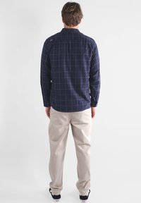 Homme portant une chemise à carreaux bleu marine à manches longues, un pantalon beige et des baskets noires et blanches, se tenant de dos contre un fond blanc.