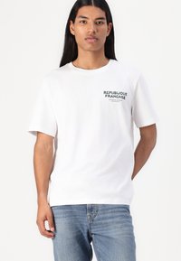 Tricou alb din bumbac, cu mâneci scurte și gât rotund. Prezintă text verde "REPUBLIQUE FRANCAISE" în zona stângă a pieptului. Croi lejer.