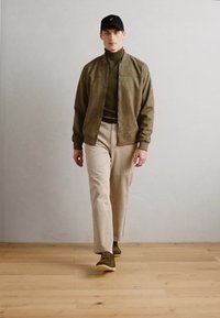 Conjunto masculino que incluye una chaqueta bomber oliva, un cuello de tortuga verde, pantalones crema y zapatillas oliva, complementado con una gorra negra.