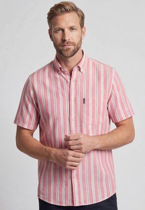 Man met baard die een kort mouw roze-wit verticaal gestreept overhemd draagt, staand tegen een effen achtergrond.