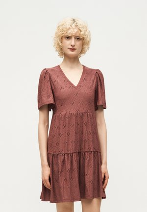 Γυναίκα με κοντά, σγουρά ξανθά μαλλιά που φοράει φόρεμα από ύφασμα με τρυπητό μοτίβο σε μοβ απόχρωση, με φουσκωτά κοντά μανίκια και V-λαιμόκοψη, στέκεται μπροστά από λευκό φόντο.