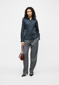 Γυναίκα με σγουρά μαλλιά που φοράει σκουρό μπλε πουκάμισο με κουμπιά, γκρι παντελόνι, μαύρες γόβες και κρατά καφέ τσάντα, στέκεται μπροστά σε λευκό φόντο.