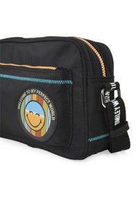 Bolso de nylon negro con un parche redondo que presenta una cara sonriente y un fondo de rayas de colores. Cierre con cremallera y costuras texturizadas en azul.