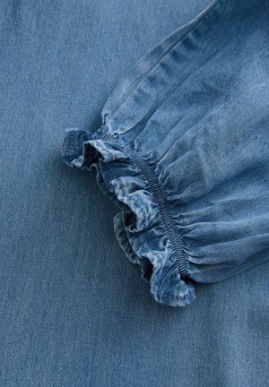 Modrá denimová tkanina předvádí shromážděný elastický manžet s volánkovými okraji, čímž vytváří texturu vedle hladkého povrchu materiálu.
