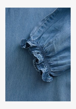 A kék farmer anyag ráncolt, gumis mandzsettát mutat be fodros szélekkel, amely textúrát teremt az anyag sima felülete mellett.