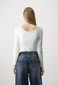 Bluză albă cu mânecă lungă și spate adânc, purtată cu blugi din denim albastru, cu talie înaltă și croială largă, având două buzunare la spate.