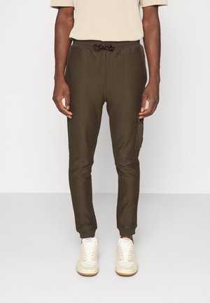 Mężczyzna noszący dopasowane ciemnooliwkowe spodnie dresowe z elastycznym ściągaczem i beżowe sneakersy, stojący na białym tle.