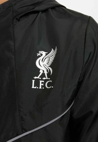 Czarna kurtka wiatrówka z białym logo Liverpool FC, gładka tekstura i odblaskowe akcenty wzdłuż szwów. Z kapturem.