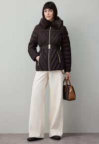 Casaco acolchoado castanho com colarinho de pelo, cintura com cinto e bolsos com fecho; combinado com calças largas creme e uma mala.